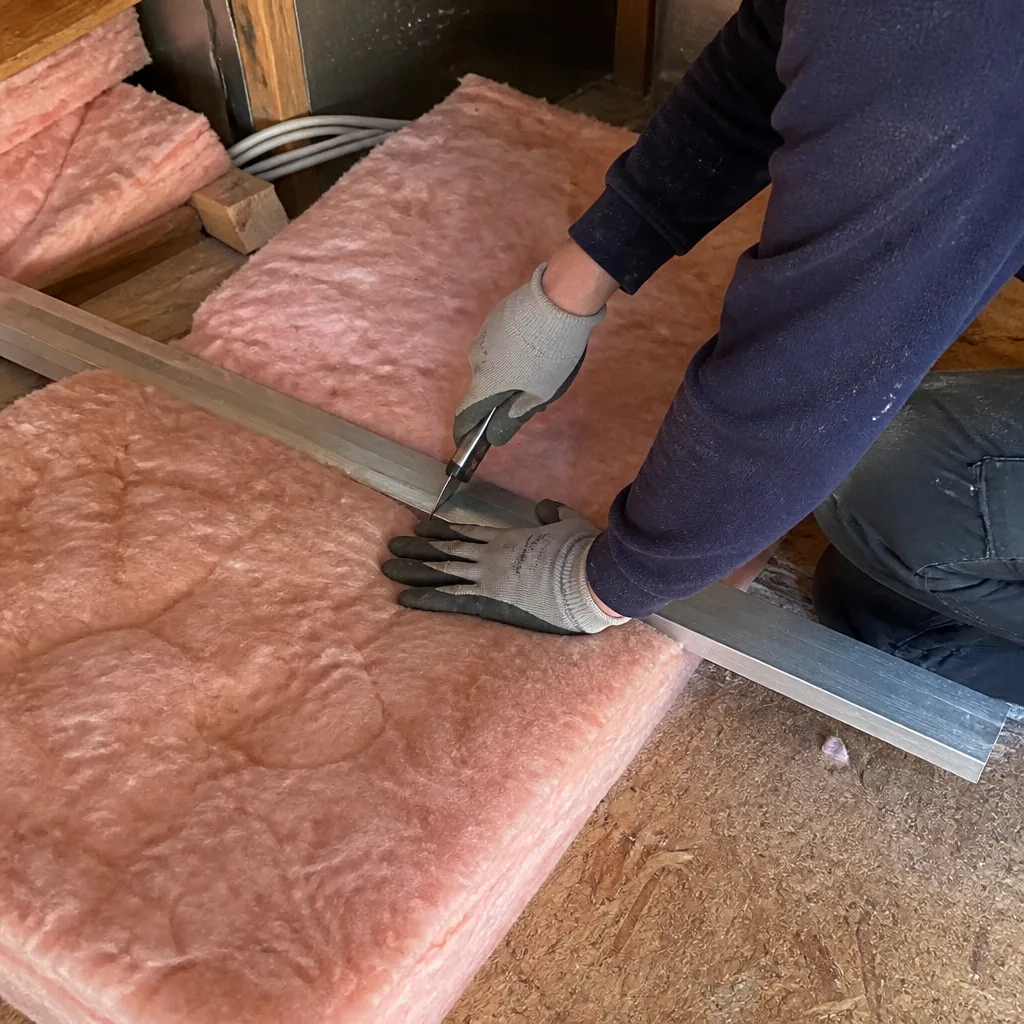 Professional contractor cutting pink fiberglass attic insulation with a utility knife.